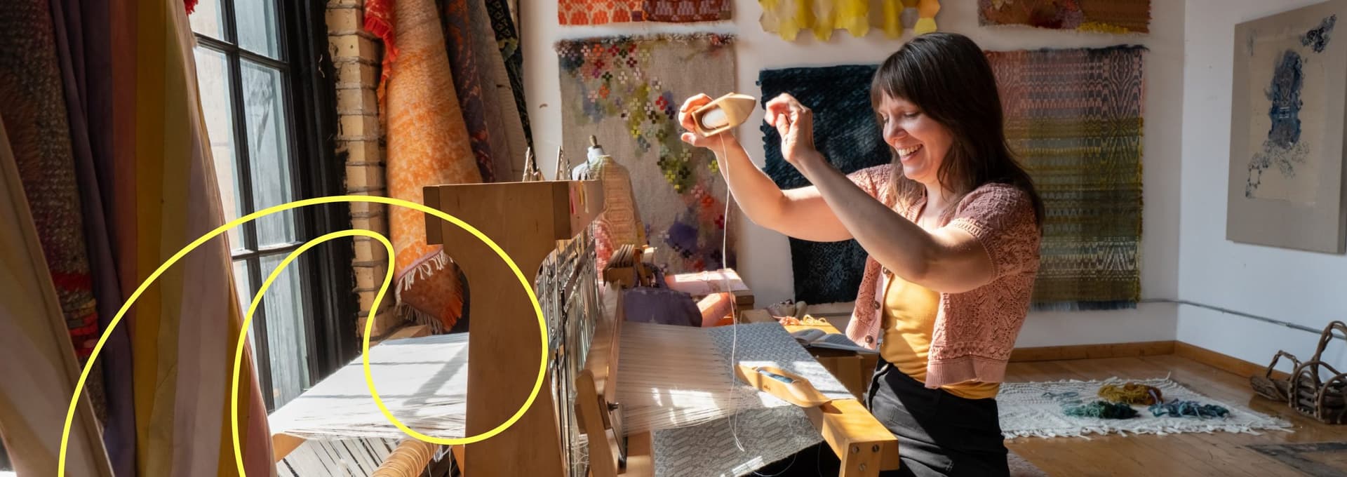 Woman working on a wooden loom, weaving fabric in a brightly lit room with textiles hanging on the walls and natural light coming through a tall window.
