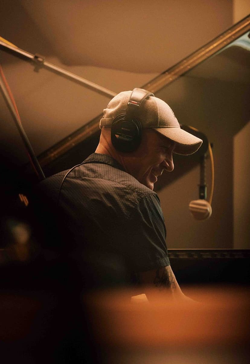Man wearing a cap and Sony headphones, smiling while playing a grand piano in a warmly lit room.
