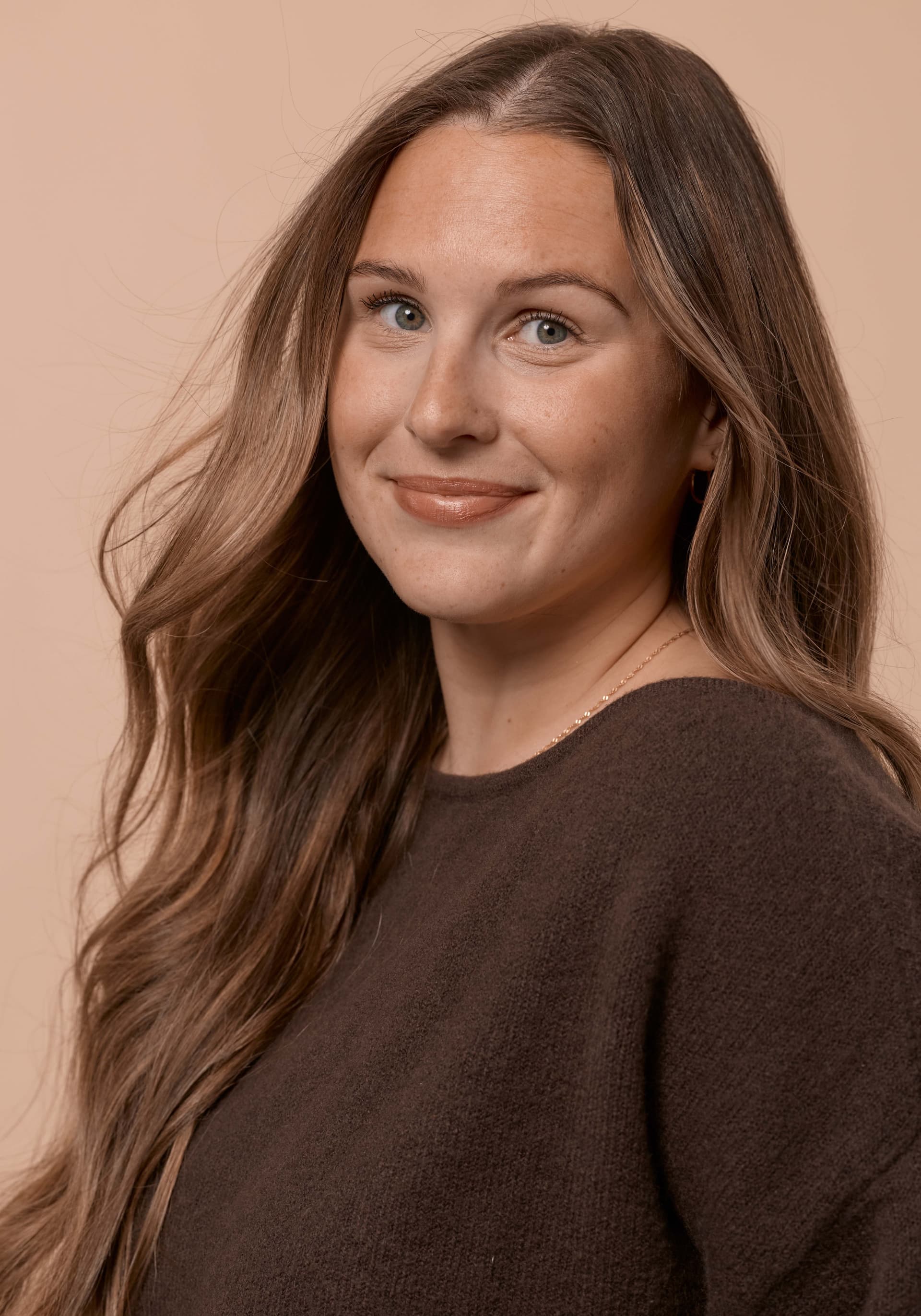 Close-up portrait of a woman with long, wavy brown hair, wearing a dark brown sweater, smiling softly against a beige background.