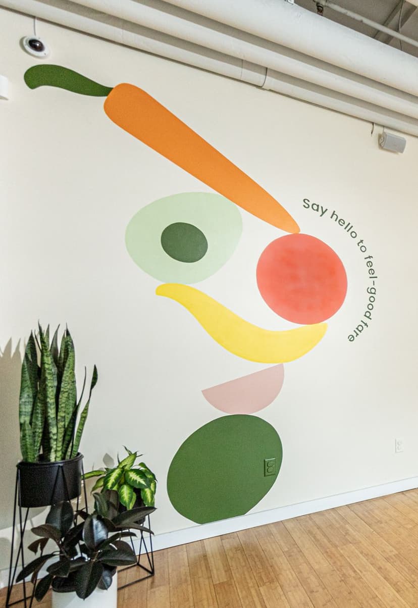 Abstract mural on white wall featuring colorful shapes resembling a carrot, avocado, banana, and other fruits or vegetables, with the phrase "say hello to feel-good fare" curved around the red circle. Several potted green plants are placed on the floor nearby.