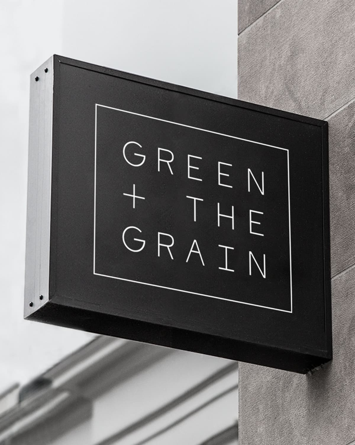 Black rectangular storefront sign with white outlined border and white text reading "GREEN + THE GRAIN," mounted on a gray wall.
