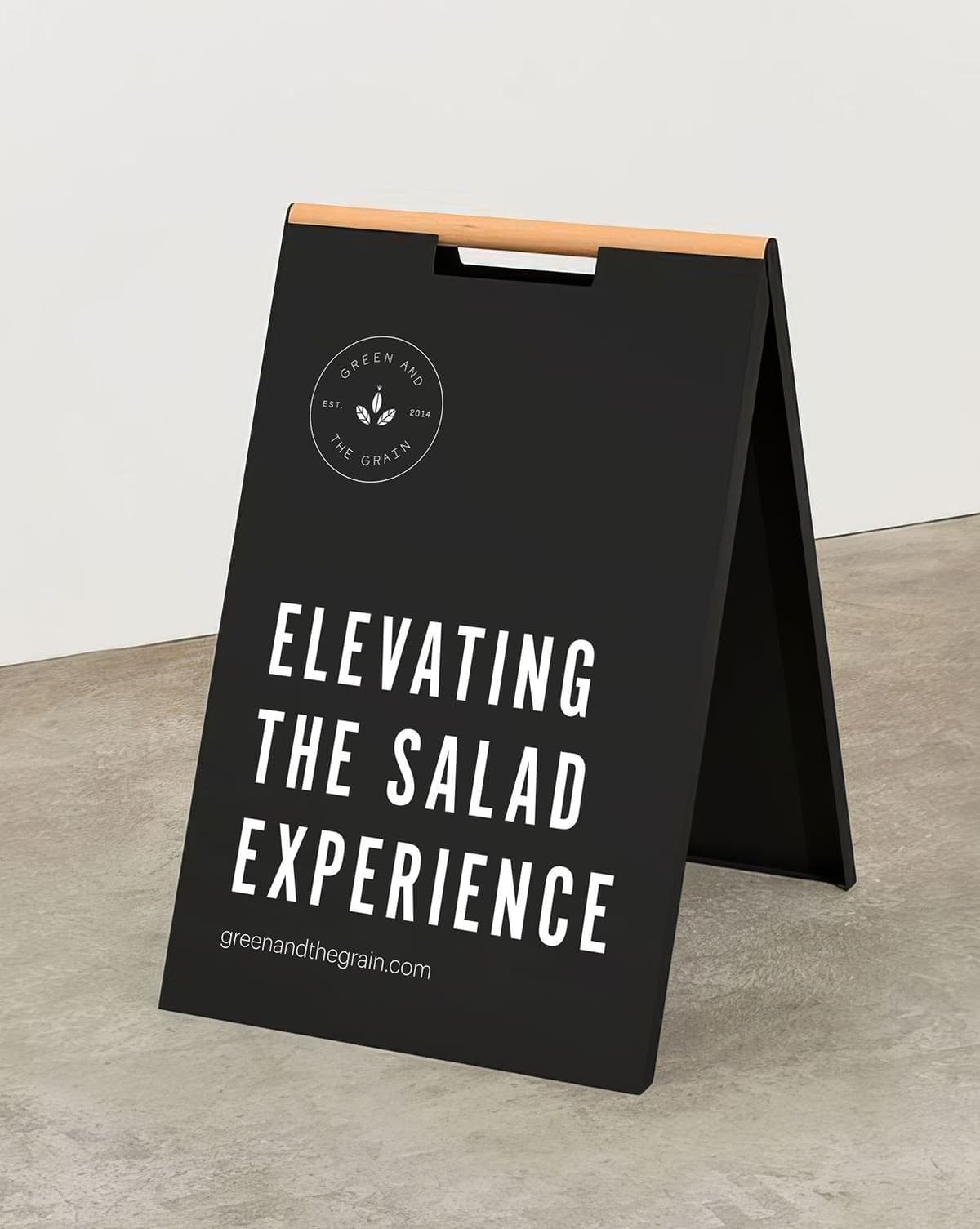 Black sandwich board sign with wooden handle and white text that reads "Elevating the Salad Experience" and a small logo above with the text "Green and The Grain Est. 2014" and the website "greenandthegrain.com" at the bottom.