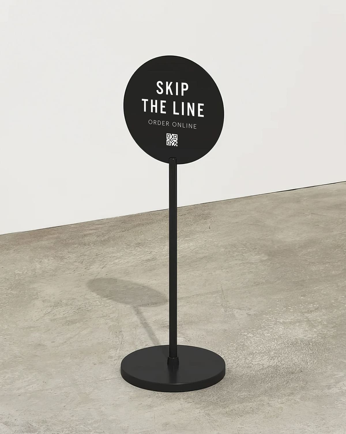 Black circular sign on a black stand with white text reading "SKIP THE LINE ORDER ONLINE" and a QR code below the text, placed on a concrete floor against a plain white wall.