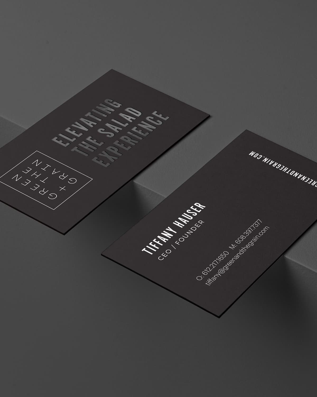 Two black business cards on a dark gray surface. One card displays the logo "Green + The Grain" and the tagline "Elevating The Salad Experience." The other card shows contact information for Tiffany Hauser, CEO and Founder, including phone numbers and an email address.