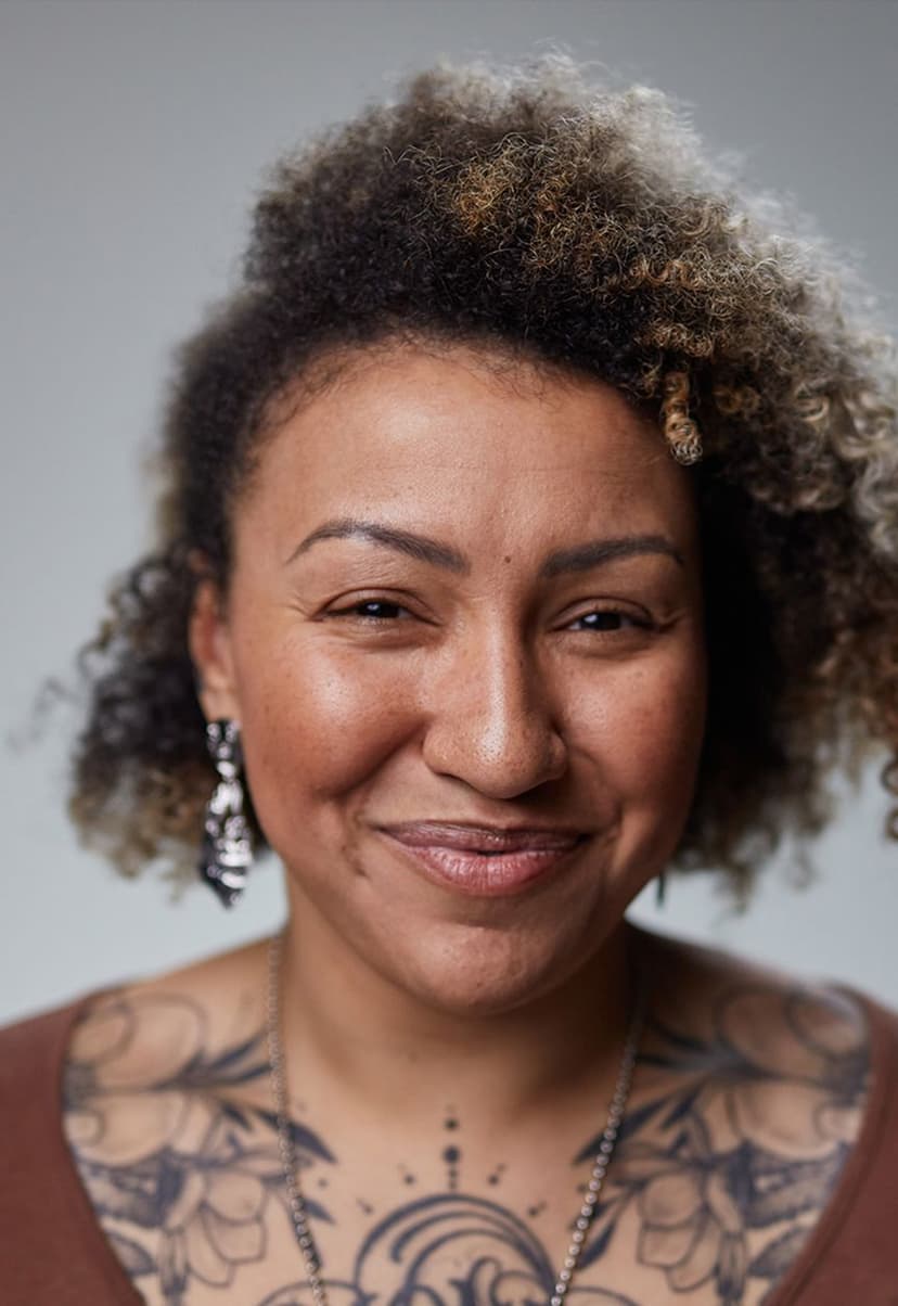 Close-up portrait of a person with curly hair, smiling gently. They have prominent tattoos on their chest and shoulders, wear dangling earrings, and a necklace against a plain gray background.