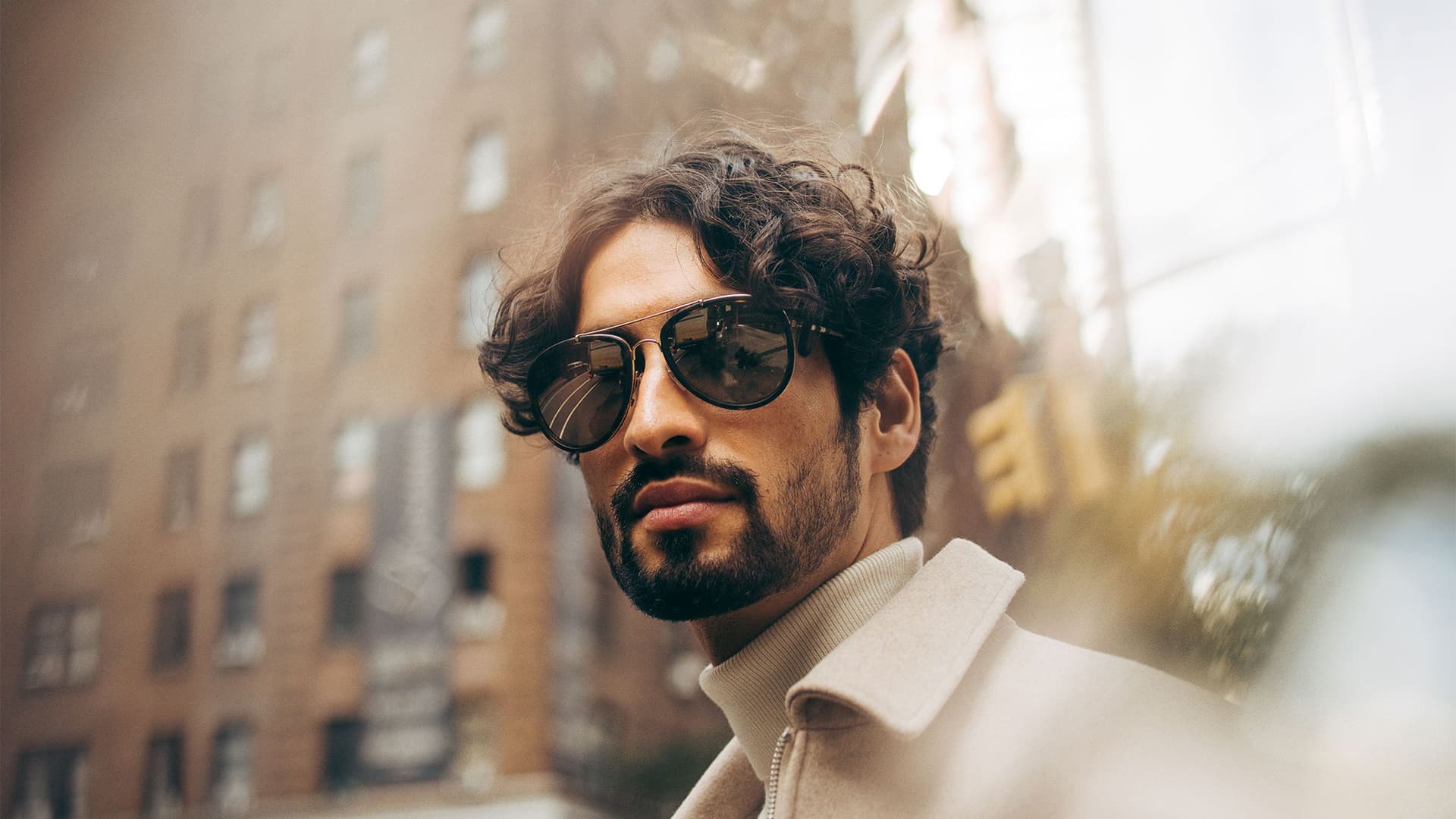Close-up portrait of a man with curly hair and a beard, wearing aviator sunglasses and a beige coat, set against a blurred city background.