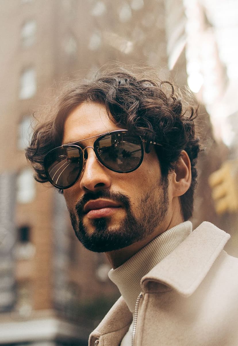 Close-up portrait of a man with curly dark hair and a beard wearing dark sunglasses, a beige coat, and a beige turtleneck sweater, with an urban city background.