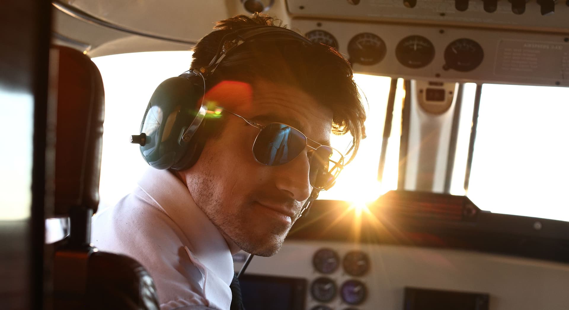 Close-up of a male pilot wearing sunglasses and a headset, seated inside a cockpit with sunlight streaming through the window behind him.