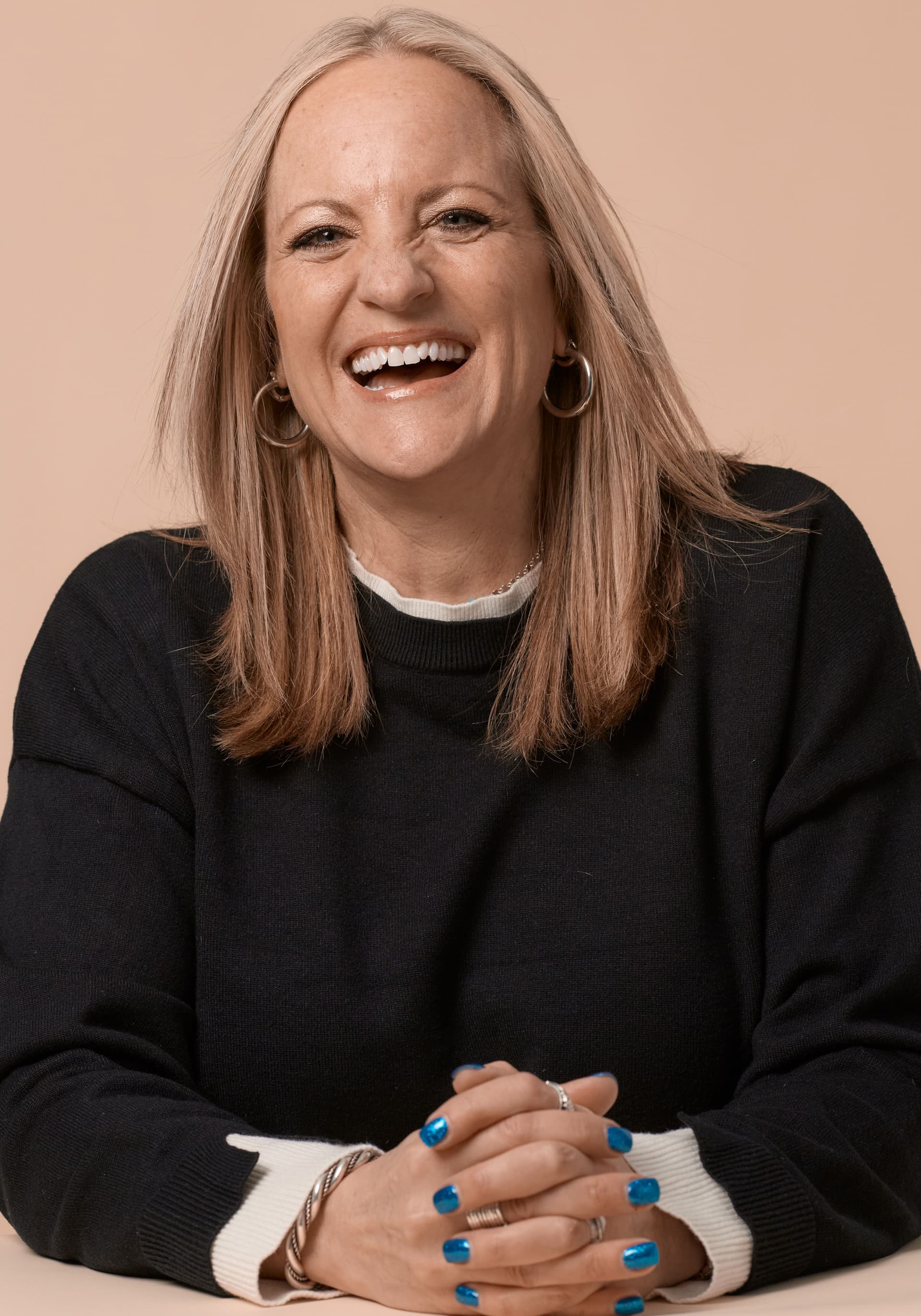 Portrait of a smiling woman with straight blonde hair wearing a black sweater, silver hoop earrings, and blue nail polish, posed with clasped hands against a beige background.