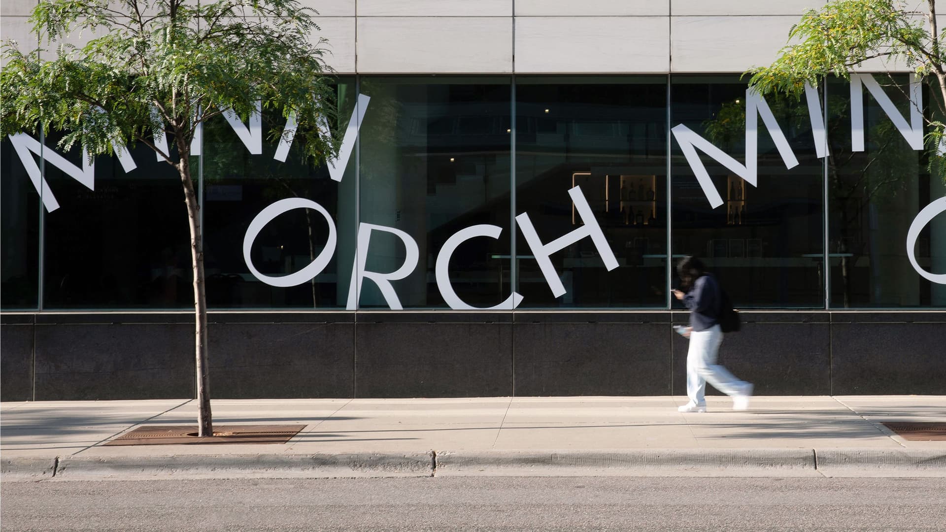 Sidewalk view of a building with large windows displaying partial curved white letters spelling "MINN ORCH MINN" and a person walking past on the sidewalk. Two small trees in grates are planted along the street in front of the building.