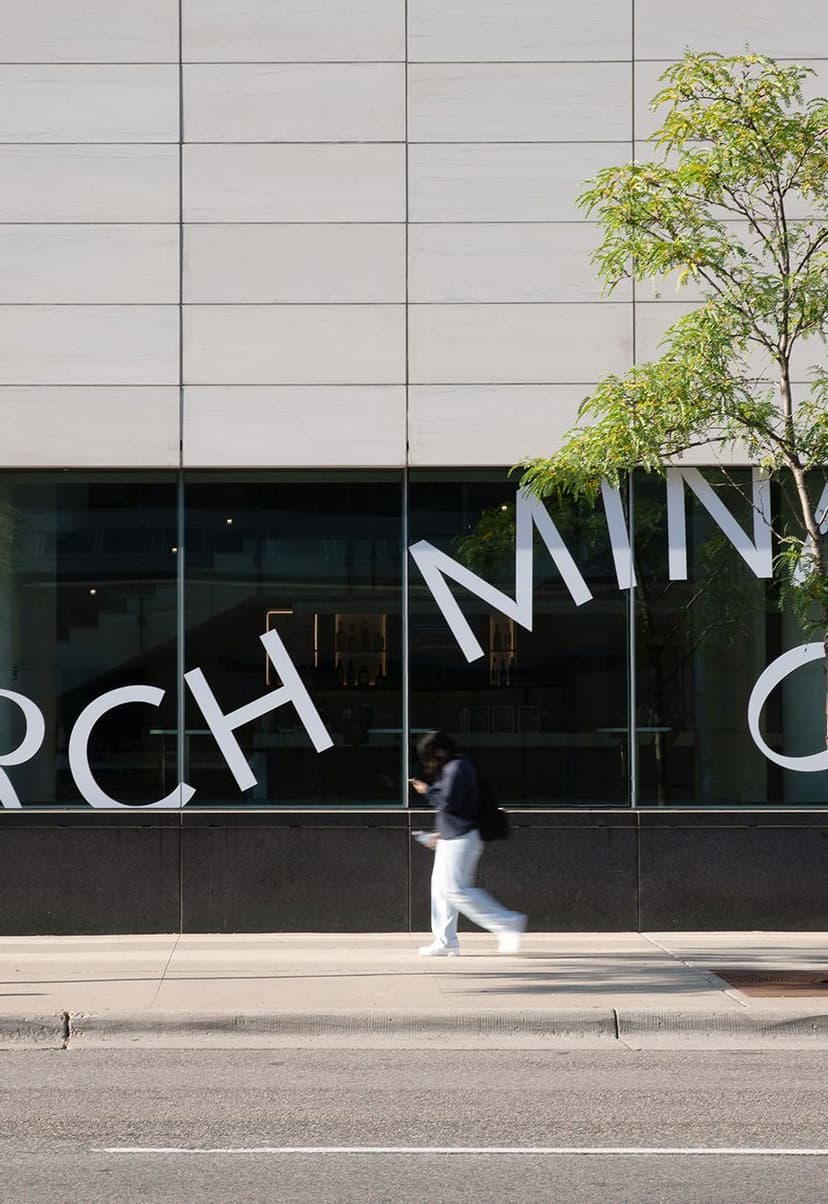 Person walking on city sidewalk in front of building with large windows displaying partial letters and a tree adjacent to the building.