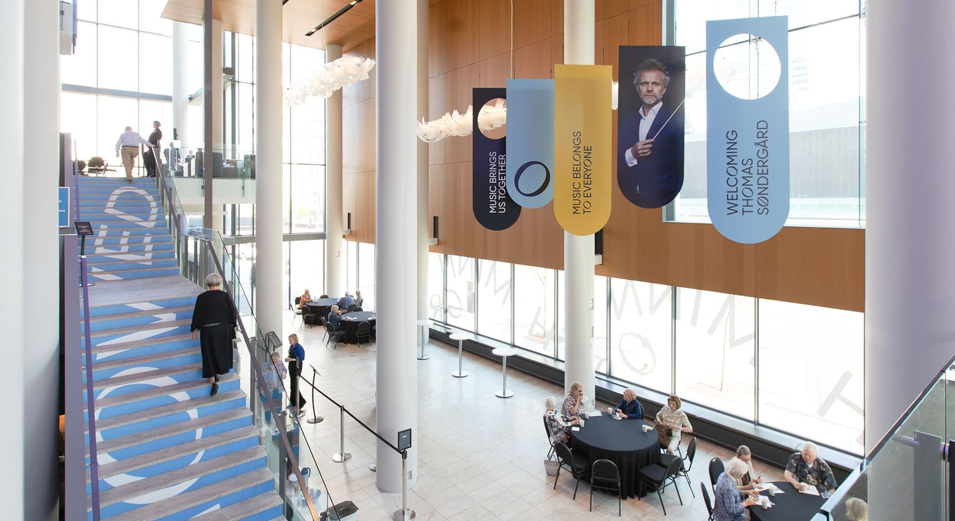 Interior of a modern building with tall white columns, large windows, and a wooden wall panel. Several people are seated at round tables covered with black tablecloths, engaged in conversation. Blue stairs on the left side have large white text. Hanging banners from the ceiling feature messages about music and an image of a man holding a conductor's baton. The banners read "MUSIC BRINGS US TOGETHER," "MUSIC BELONGS TO EVERYONE," and "WELCOMING THOMAS SØNDERGÅRD."
