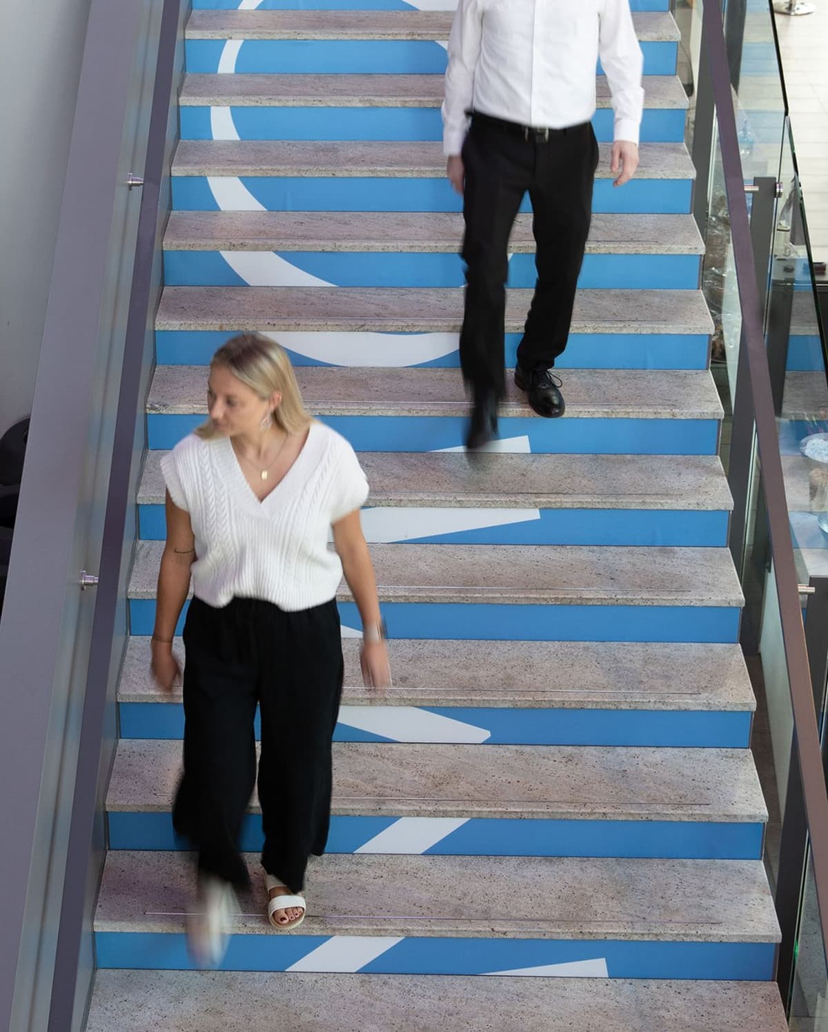 Two people walking on a stairway with blue risers featuring white arrow designs; a woman wearing a white sleeveless top and black pants is walking down, and a man in a white shirt and black pants is walking up.