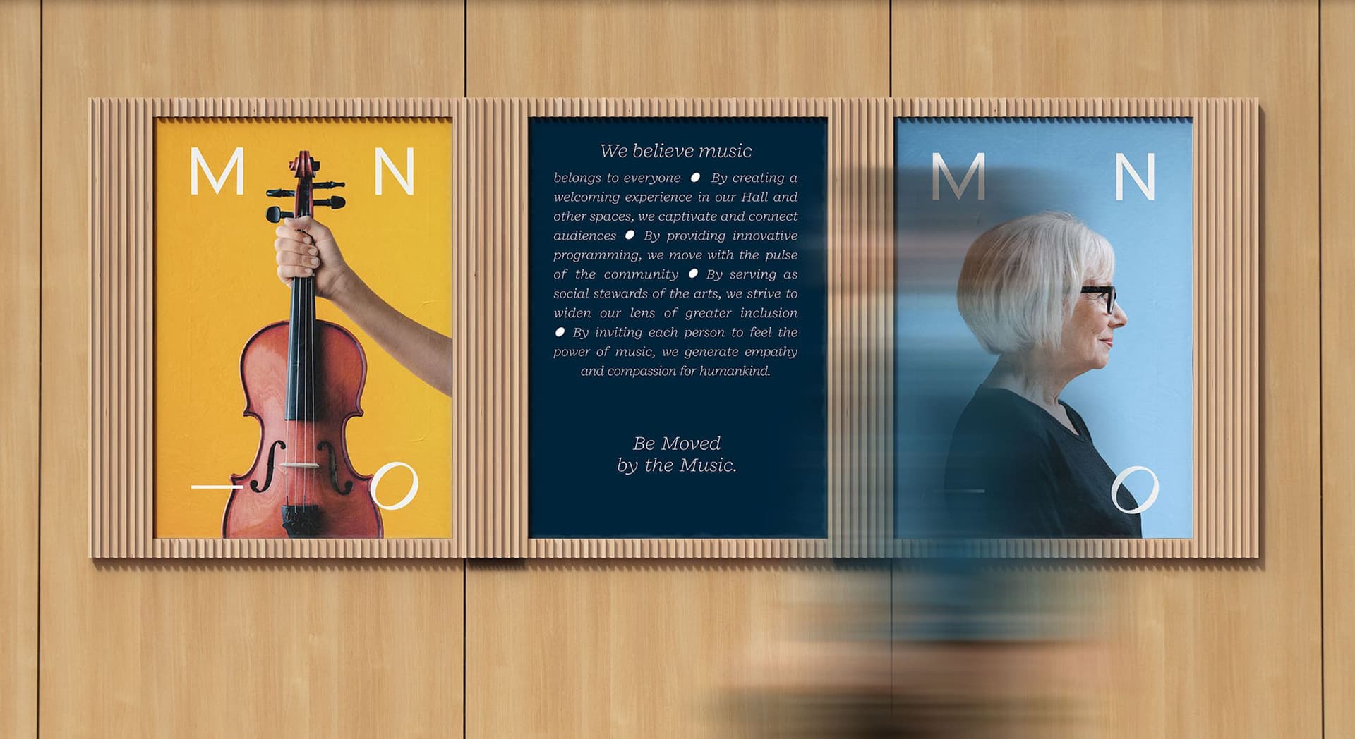 Triptych posters mounted on a light wood wall. The left poster features a hand holding a violin against a yellow background with letters "M N - O." The center poster has a navy blue background with text about music's inclusivity and impact, ending with "Be Moved by the Music." The right poster shows a profile of a woman with short white hair and glasses against a blue background with letters "M N - O." A blurred person walks past in front of the posters.
