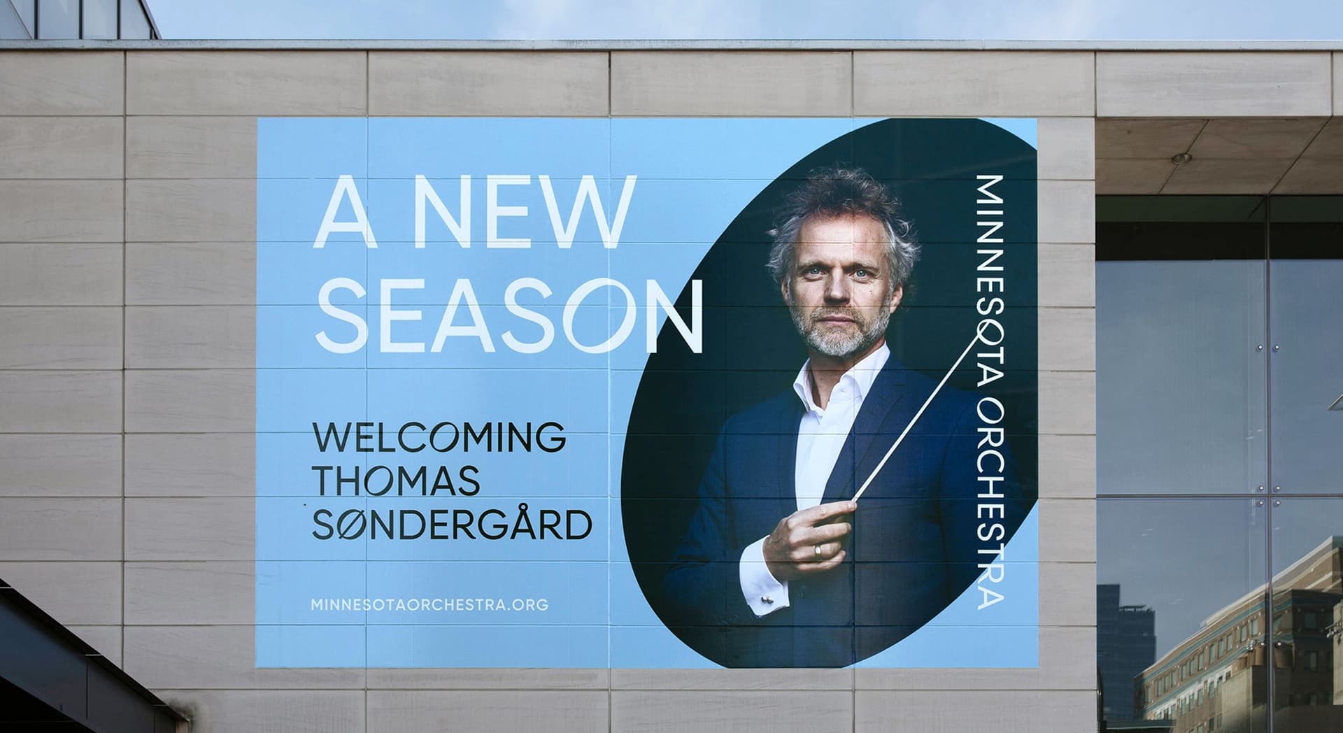 Outdoor billboard on a building wall showing a man in a blue suit holding a conductor's baton. The text reads, "A New Season Welcoming Thomas Søndergård," and "Minnesota Orchestra" along with the website "minnesotaorchestra.org."