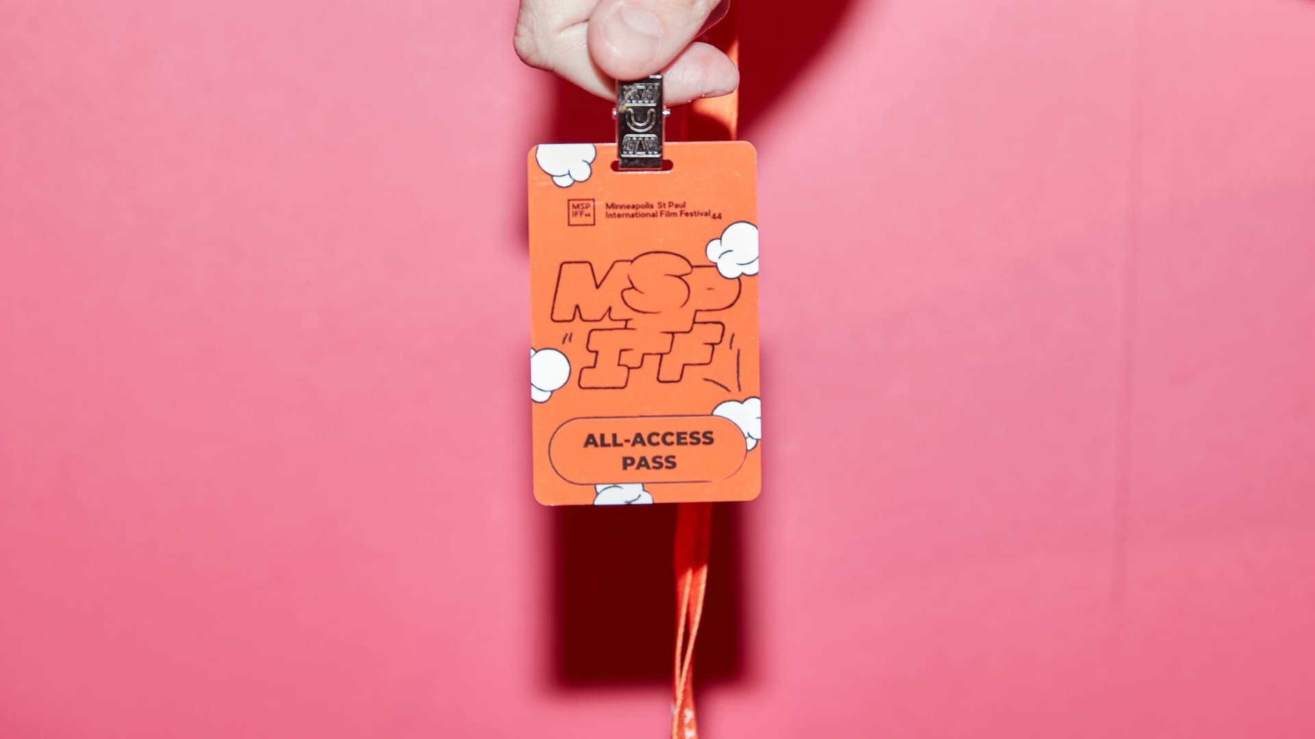 Hand holding an orange Minneapolis St Paul International Film Festival all-access pass against a pink background.