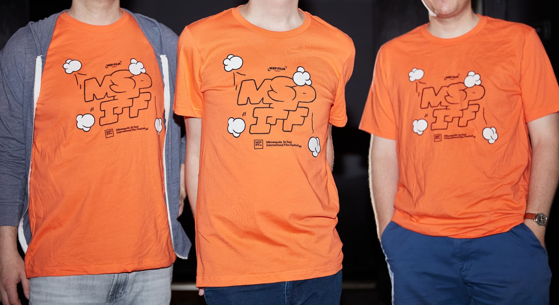 Three people standing side by side wearing bright orange T-shirts with a cartoonish design and the text "MSP IFF" and "Minneapolis St Paul International Film Festival 44" printed on them. The person on the left also wears a gray zip-up hoodie, the person in the middle wears dark jeans, and the person on the right wears blue pants with a watch on the left wrist. The background is dark and out of focus.