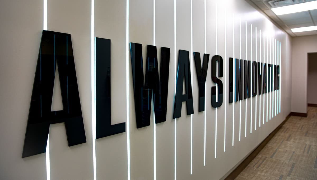 Black 3D letters mounted on a beige wall spell out "ALWAYS INNOVATIVE," with vertical white LED light strips between each letter, creating a modern and sleek appearance in a hallway with carpeted floor and ceiling lights.