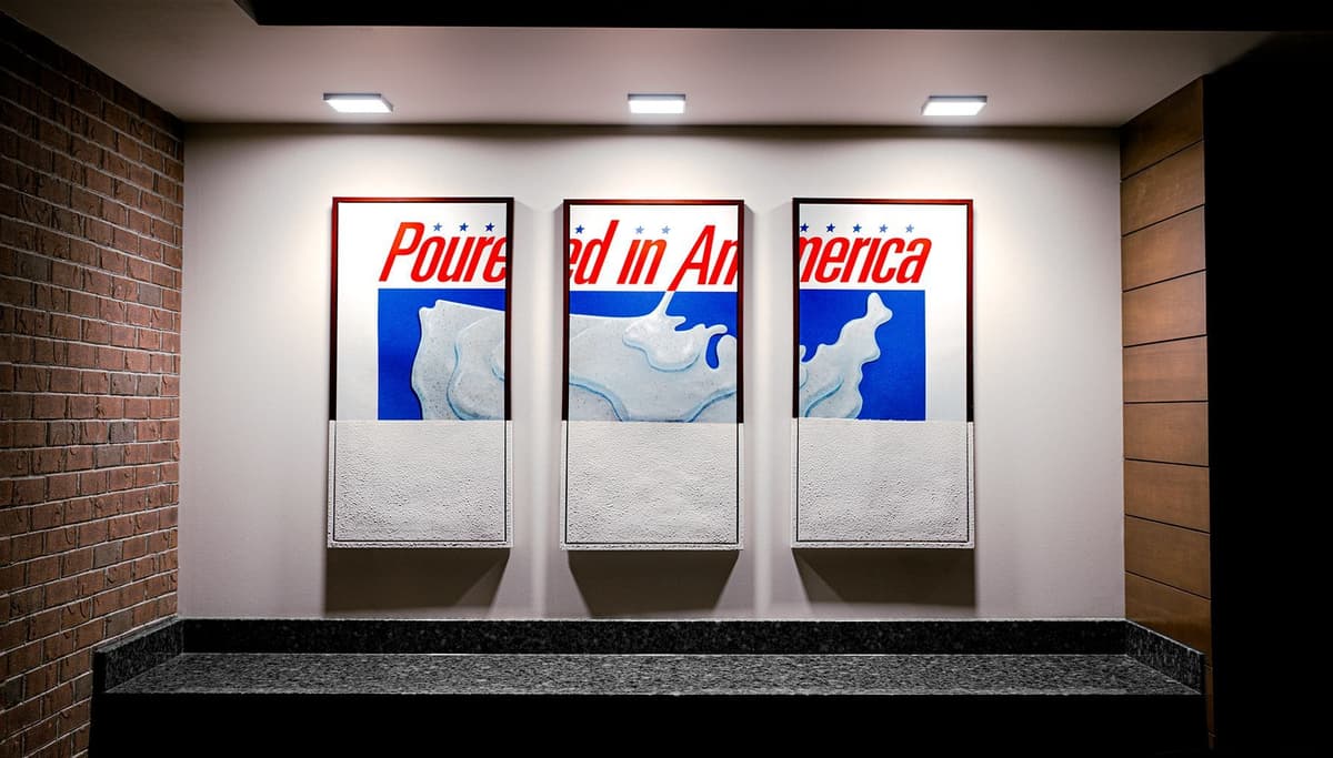 Triptych wall art featuring a stylized map of the United States with a splash of milk and the partially visible red text "Poured in America" set against white and blue backgrounds. The panels are mounted on a white wall, illuminated by three ceiling lights, with a black granite surface beneath and brick and wood-paneled walls on each side.