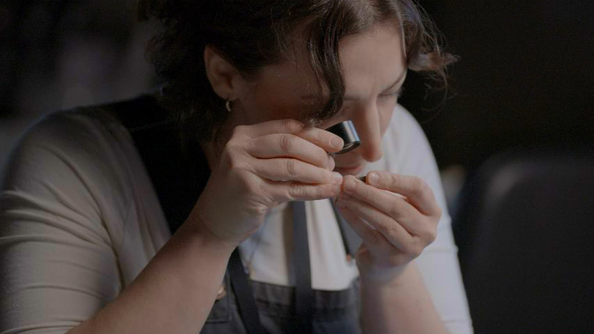 Person closely examining a small object with a magnifying loupe, wearing a white shirt and a dark apron, against a dark background.