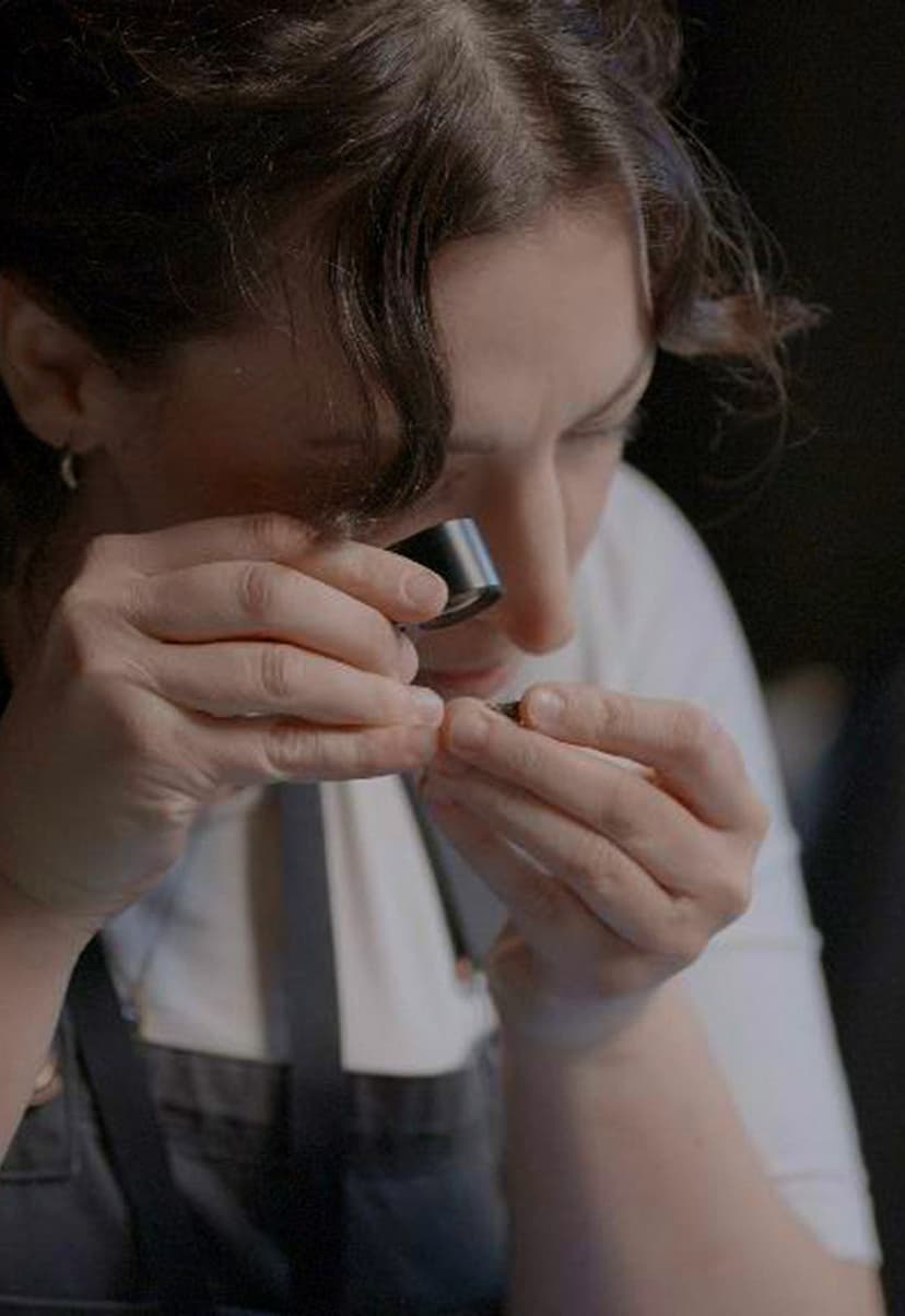 Close-up of a person closely examining a small object using a jeweler's loupe, wearing a white shirt and dark apron.