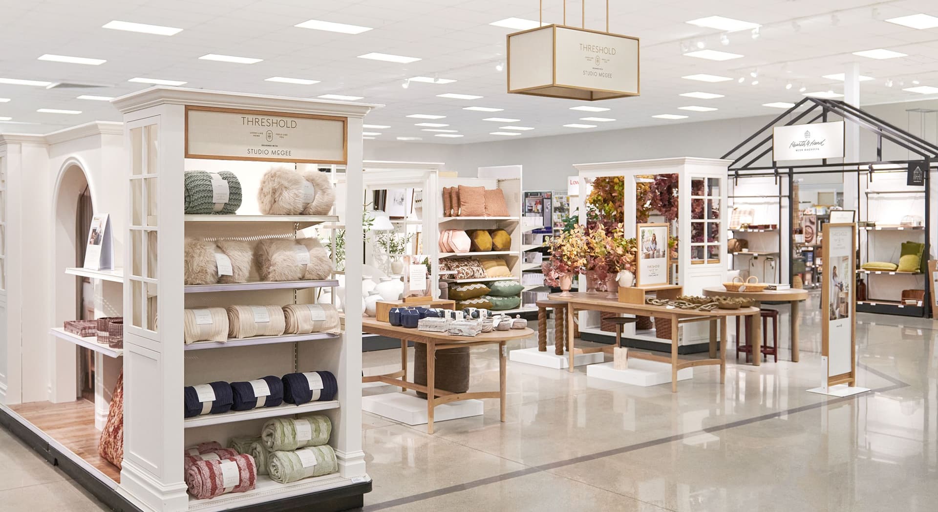 Retail display area featuring home decor items including blankets, pillows, vases, and artificial plants. Shelves and tables are arranged neatly with various textures and colors in a bright, spacious store interior under fluorescent lighting. Signs indicate brands "Threshold Studio McGee" and "Hearth & Hand with Magnolia."