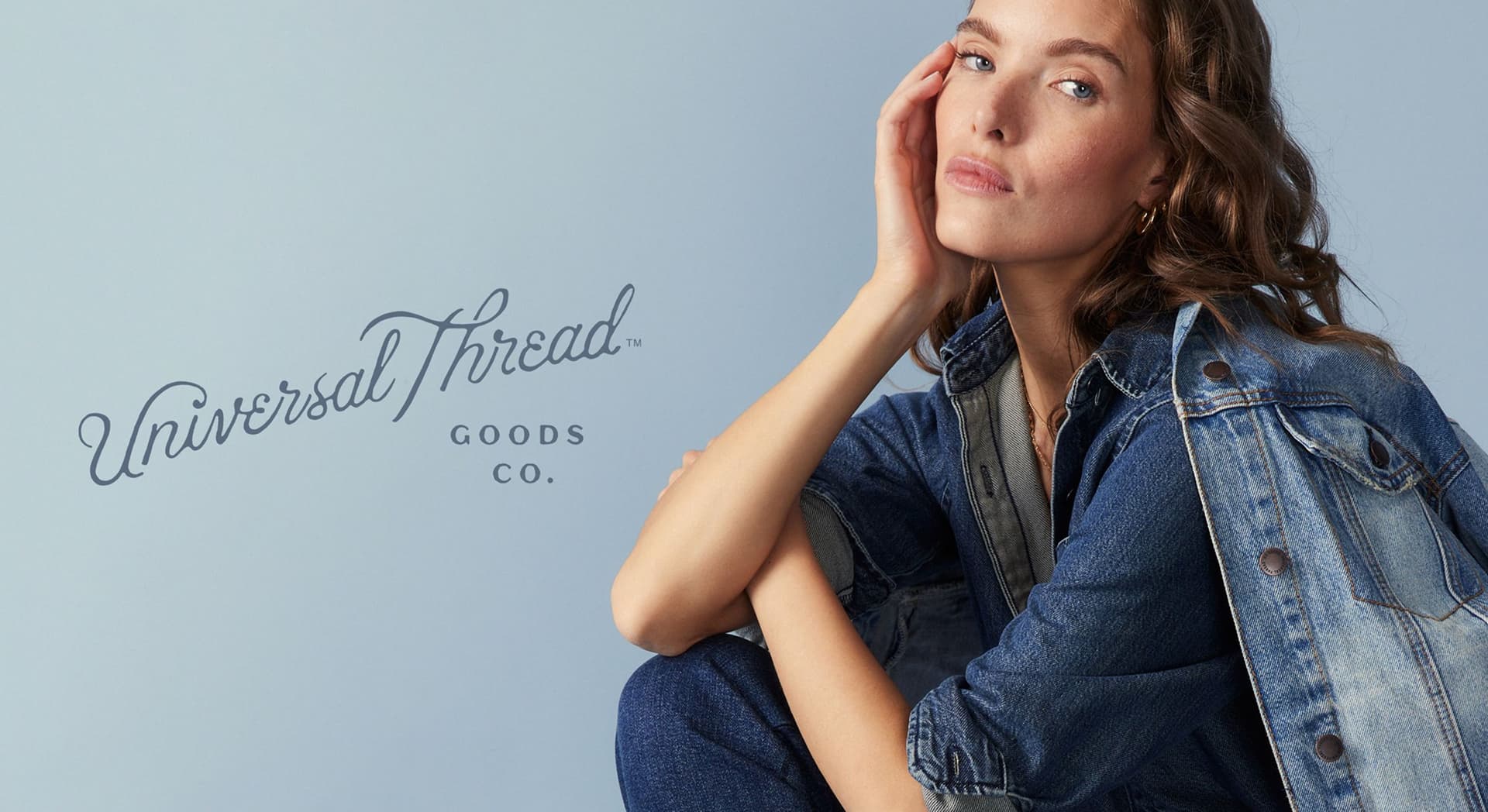 Woman with wavy brown hair wearing denim jacket and jeans, sitting with her hand resting on her face against a light blue background with the text "Universal Thread Goods Co."