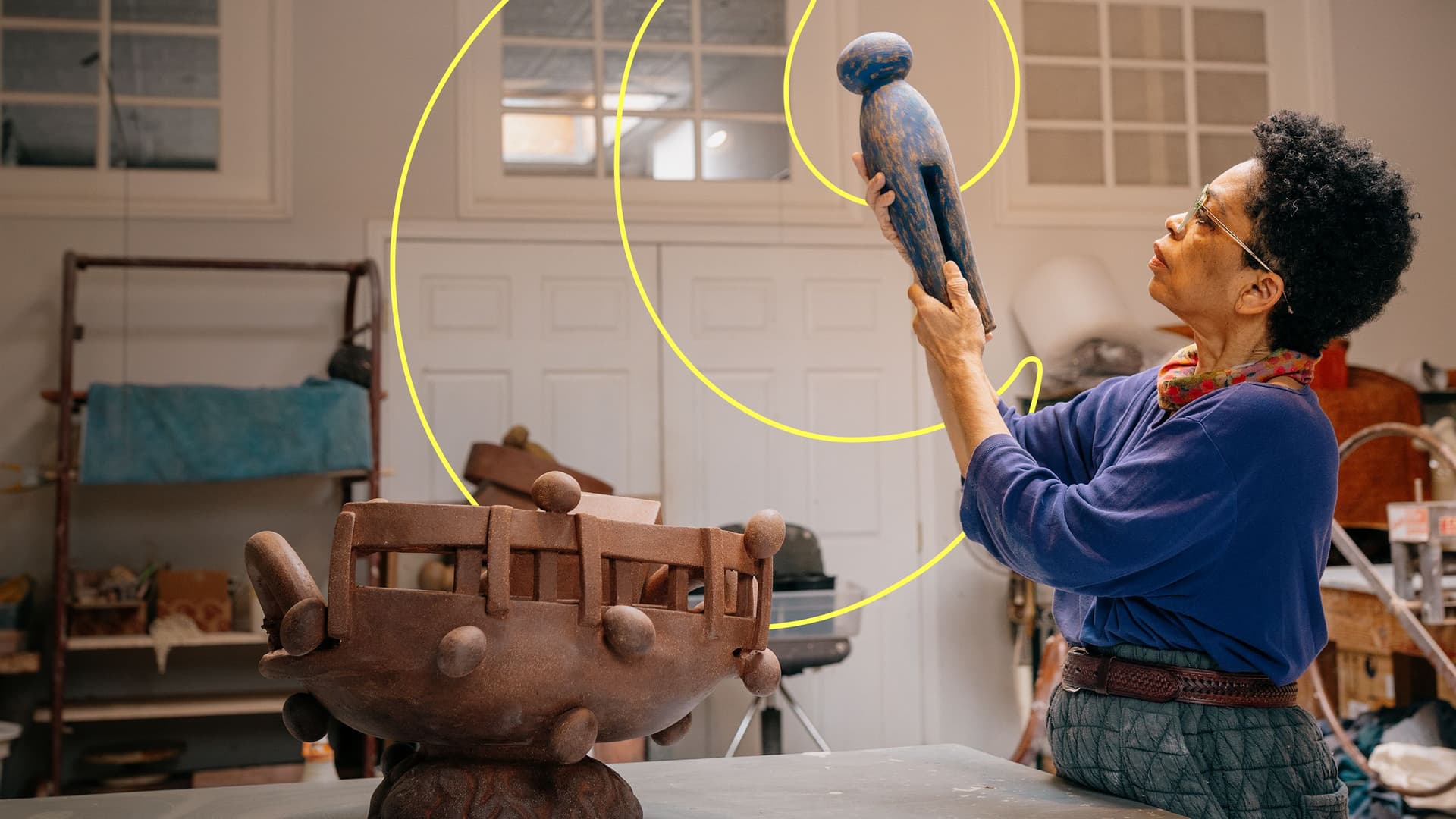 Woman with short curly hair and glasses, wearing a purple top and scarf, holding up and examining a blue wooden sculpture of a human figure in an art studio with other art pieces and art supplies.