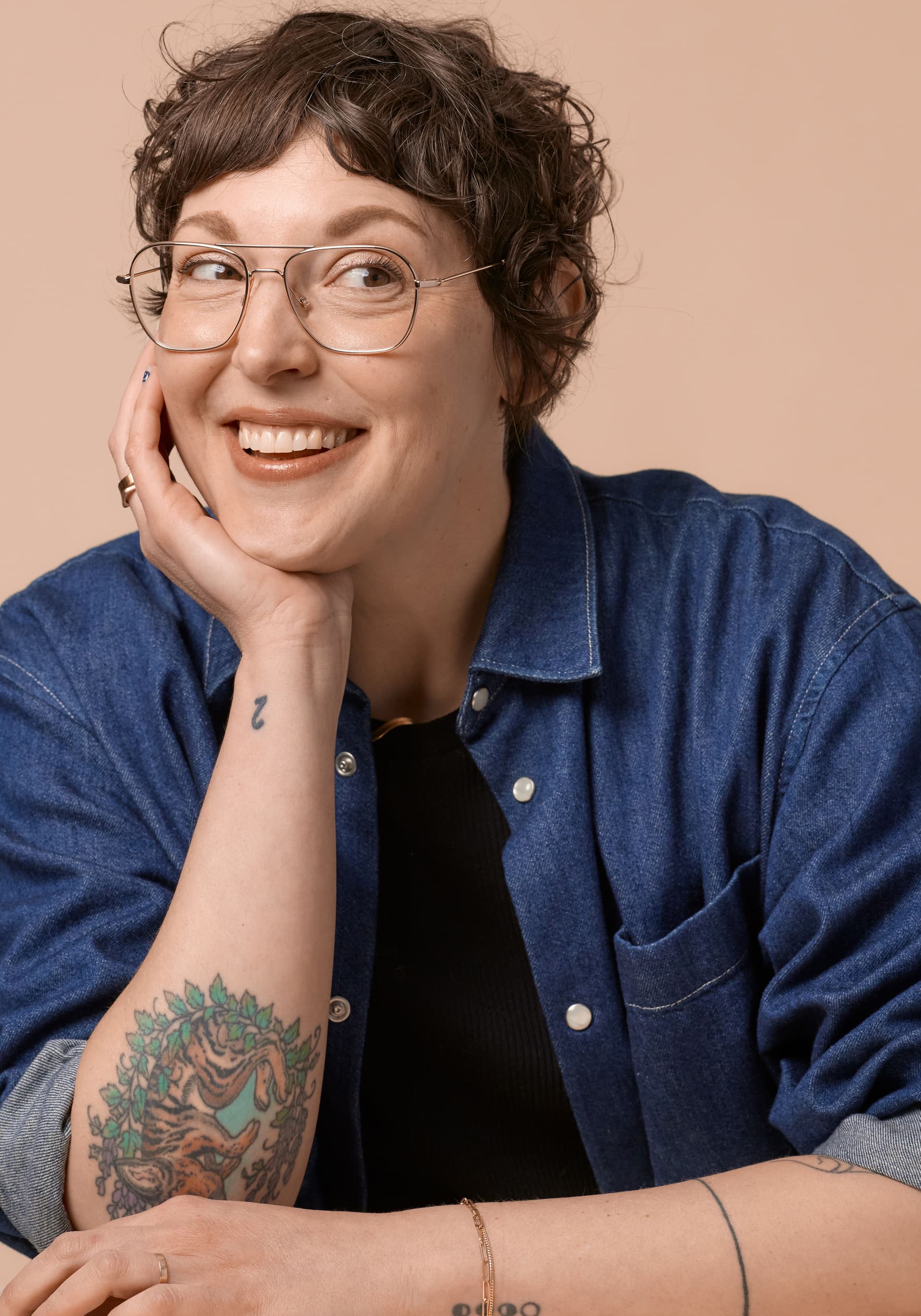 Smiling person with short curly hair and glasses, resting their head on their hand, wearing a blue denim shirt over a black top, with visible tattoos on their forearm and wrist against a beige background.