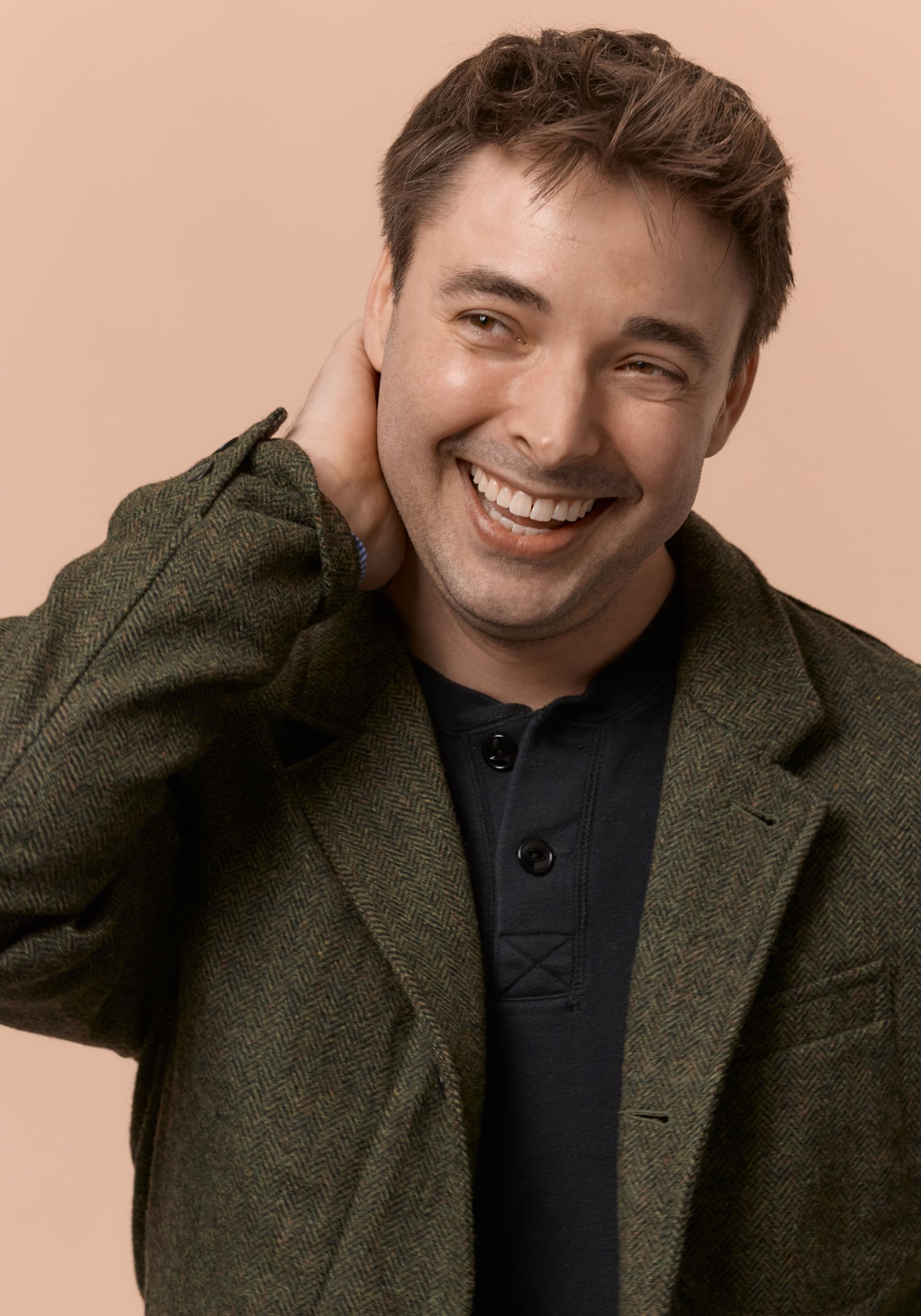 Smiling man with short brown hair wearing a green tweed jacket and black shirt, touching the back of his neck against a beige background.