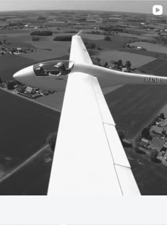 Black and white aerial photo of a glider aircraft flying over a rural landscape with fields and scattered buildings.
