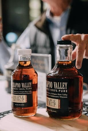 Two bottles of Kakapoo Valley 100% Pure Maple Syrup on a table, one smaller and one larger, with a person reaching for the larger bottle in the background.