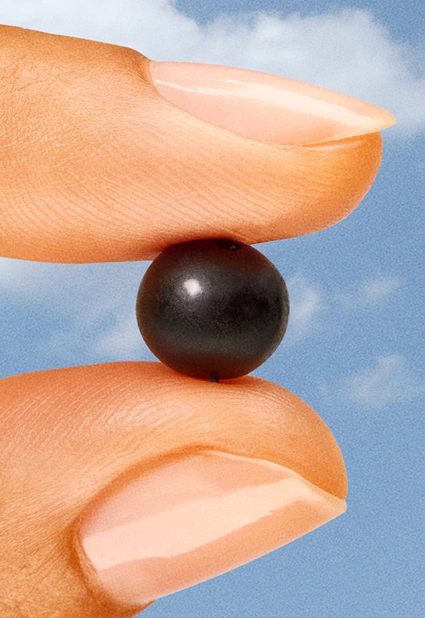 Close-up of a hand with manicured nails holding a small, round, shiny black berry between the thumb and index finger against a blue sky background with soft clouds.