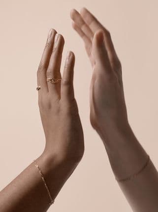 Two raised hands facing each other against a neutral background, one adorned with delicate gold rings and a bracelet.