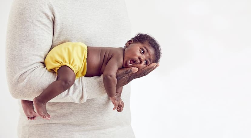 Adult wearing a light gray sweater holding a baby in yellow shorts lying face down on their arm against a plain white background. The baby has an open mouth and wide eyes.