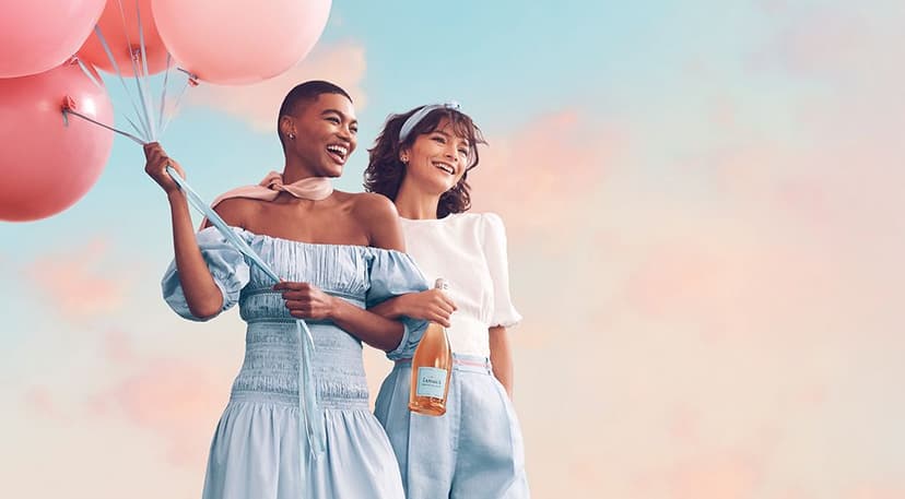 Two smiling women stand close together against a pastel sky background; the woman on the left holds several pink balloons and wears a light blue off-shoulder dress, while the woman on the right holds a bottle of sparkling beverage and wears a white blouse with light blue pants.