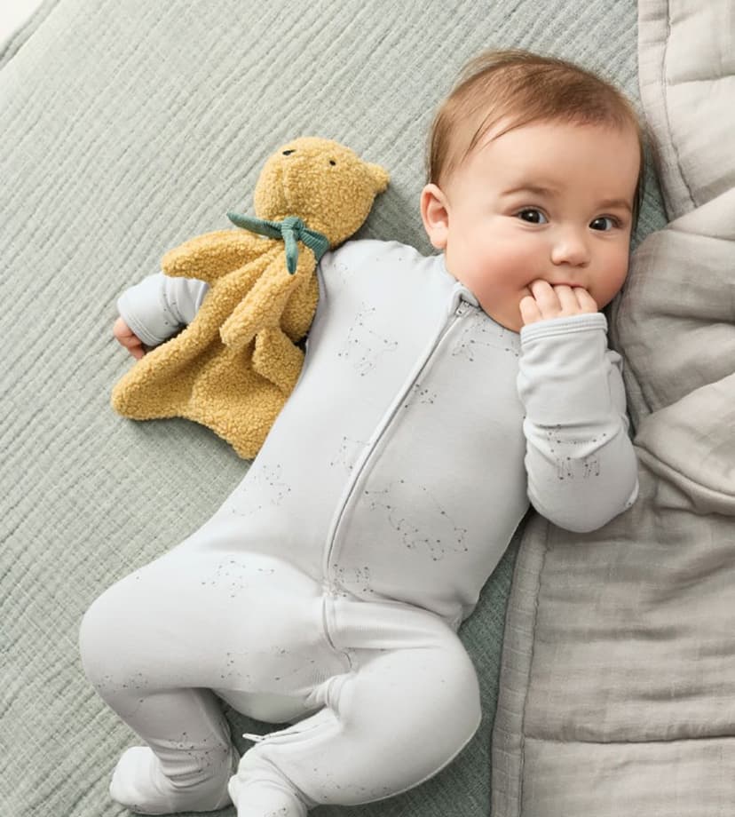 Baby in a light grey sleeper lying on a grey quilted blanket with a small tan plush bear blanket beside them, with fingers in their mouth.