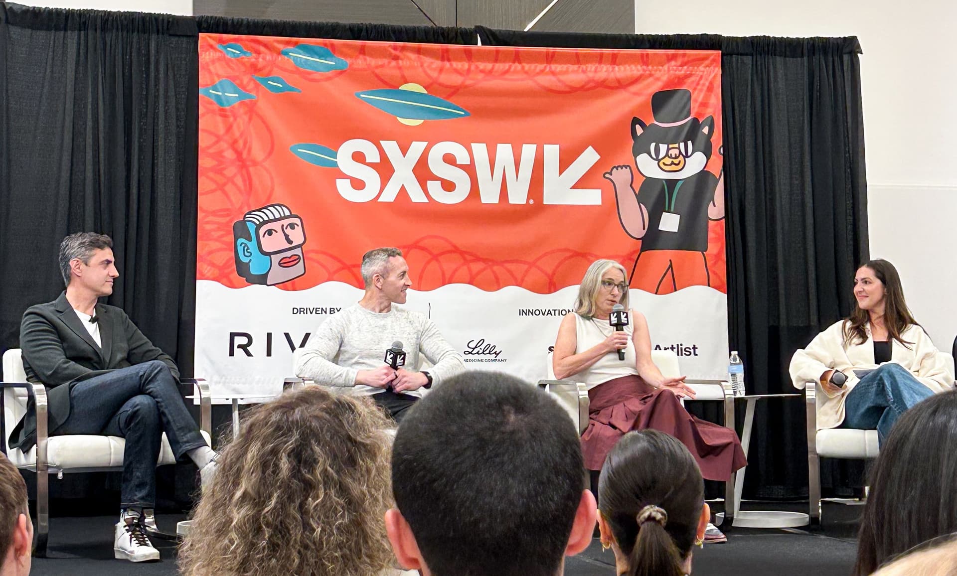 Panel discussion at SXSW event with four speakers seated on stage holding microphones, a large orange SXSW banner with illustrations behind them, and audience heads visible in the foreground.