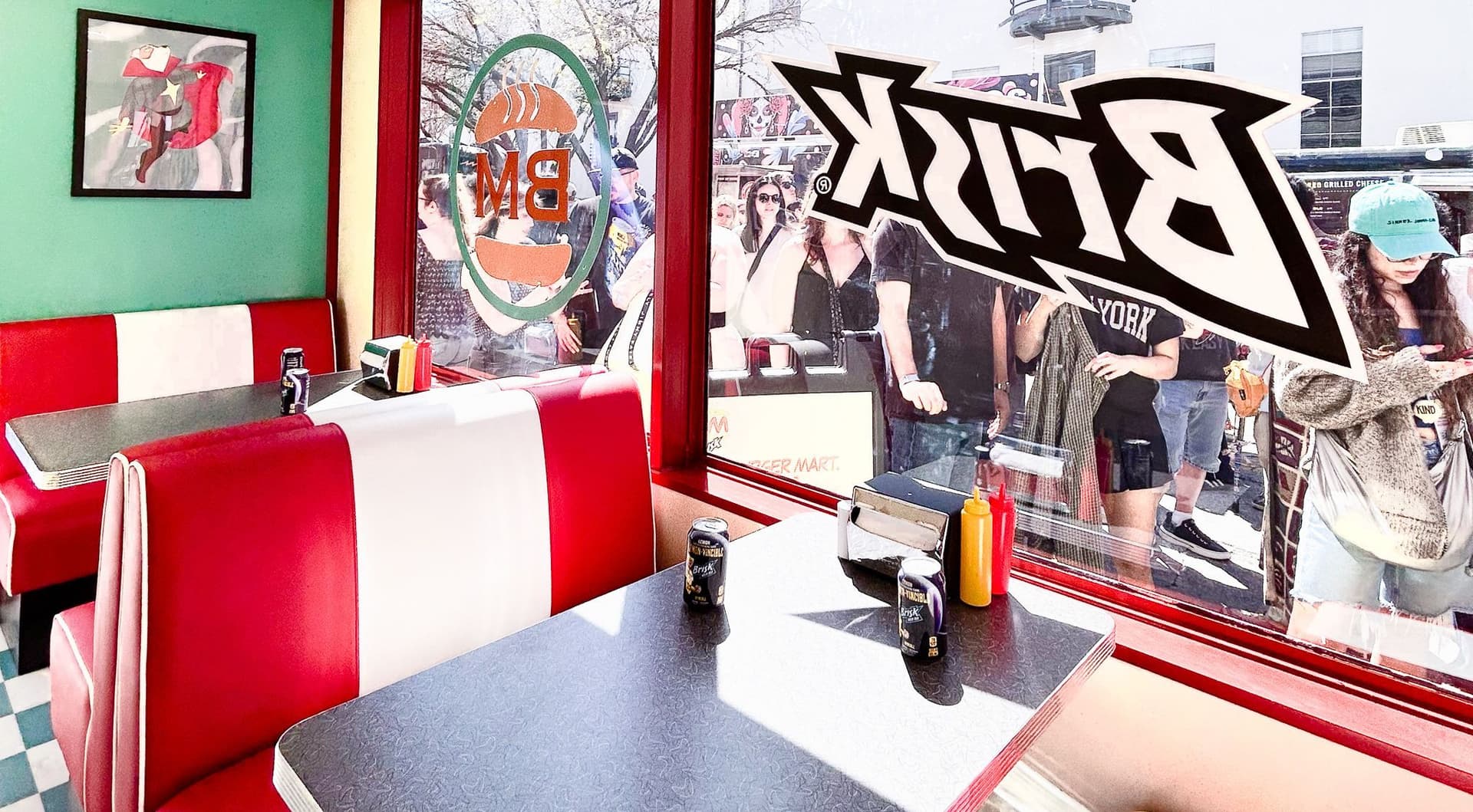 Interior of a retro diner with red and white booth seating, gray laminate tables, and condiments on the tables. A large window shows people waiting outside, with the reversed "Brisk" logo decal on the glass. A framed abstract artwork is on the mint green wall.