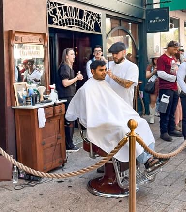 Barber giving a haircut to a seated customer outside a vintage-style barbershop, with people standing and watching nearby on a city sidewalk.
