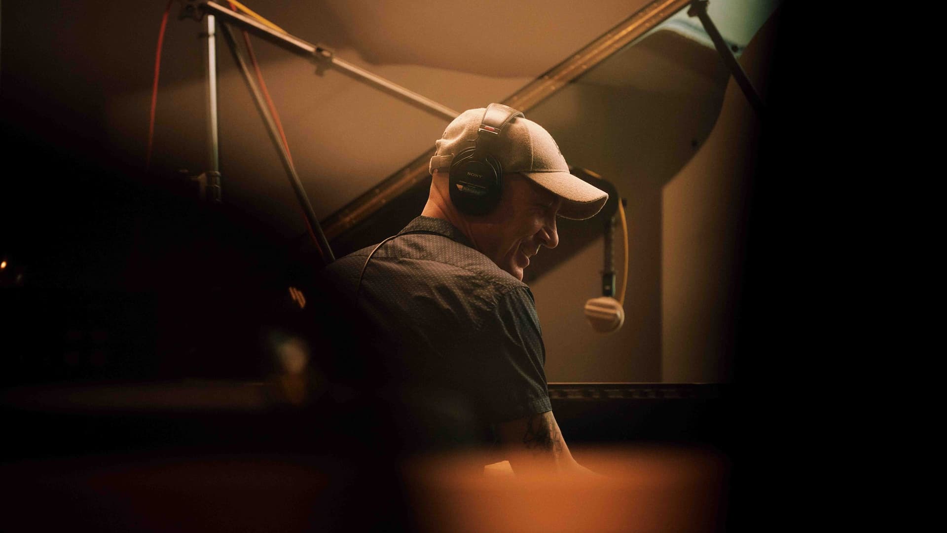 Man wearing a beige cap and headphones, smiling while playing a piano in a dimly lit room with a microphone suspended above.