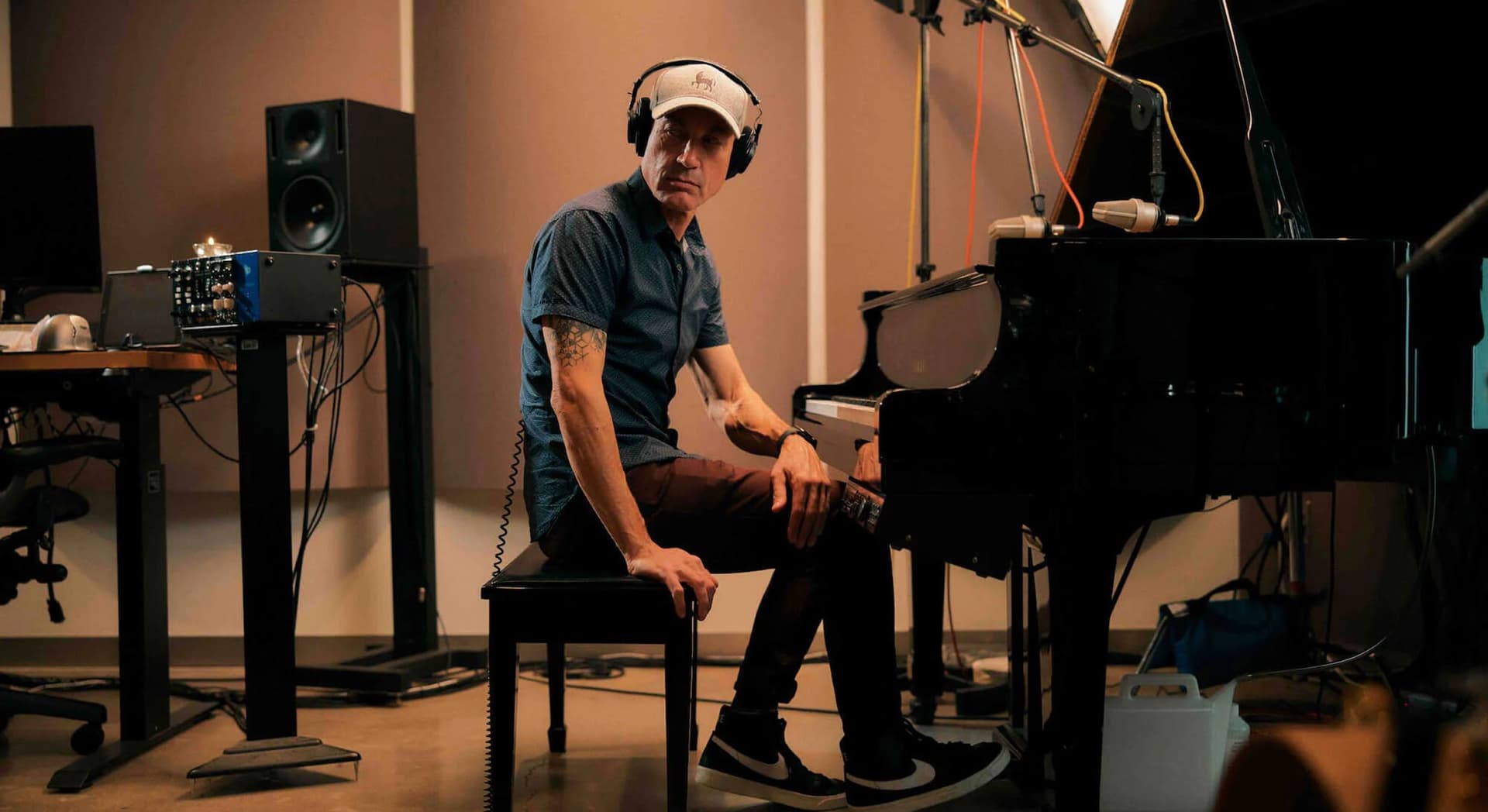 Man wearing headphones and a cap, sitting on a piano bench in a recording studio, with a grand piano and audio equipment around him.