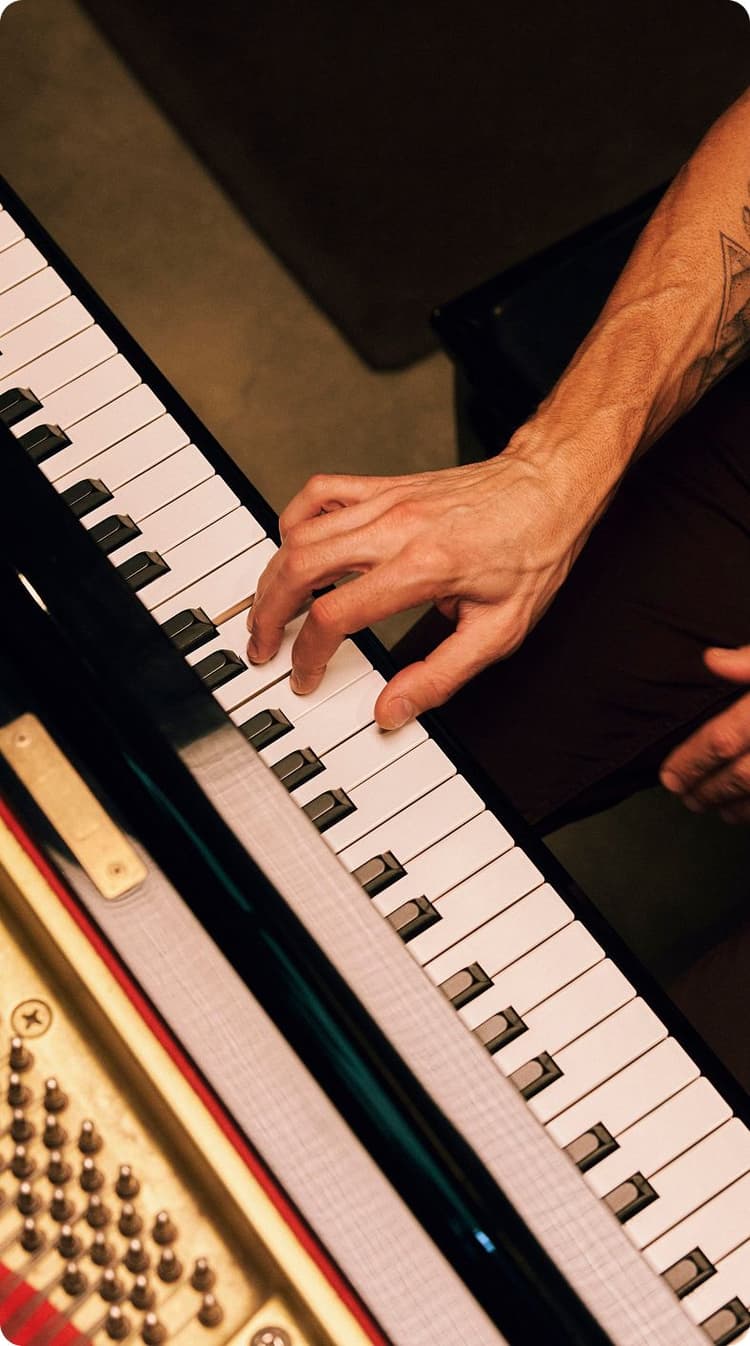 Close-up of a person's left hand playing keys on a grand piano, showing the piano strings and tuning pins.