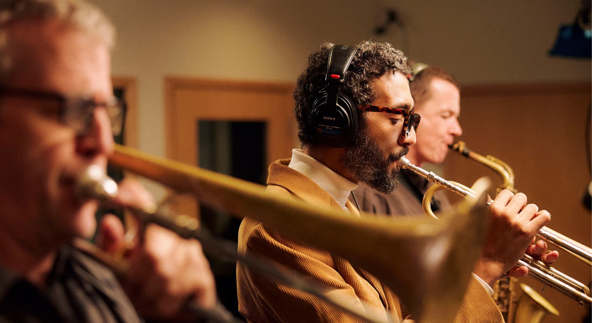 Three male musicians playing brass instruments in a recording studio, one wearing headphones and glasses, focused on their performance.