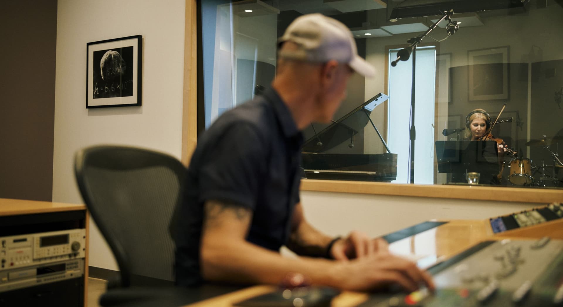 Man wearing a cap operates sound mixing equipment in a recording studio control room, while a woman wearing headphones plays violin inside the soundproof recording booth.