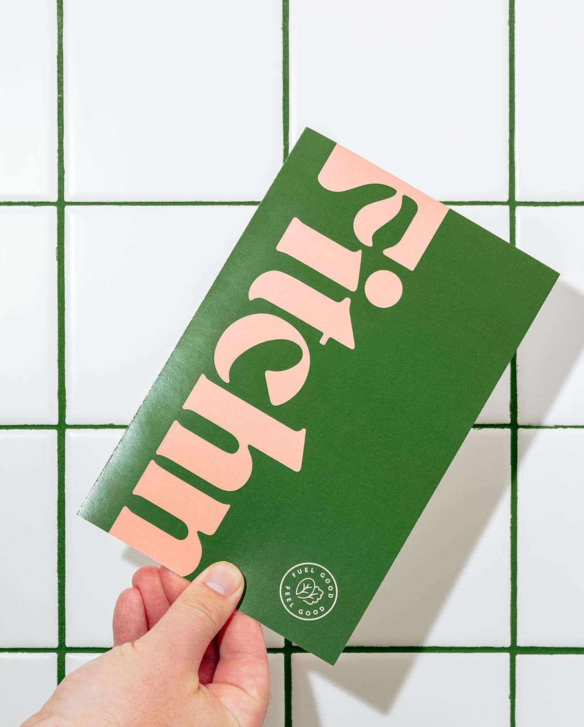 Hand holding a green card with large pink text, partially visible "yicum," and a circular logo at the bottom right corner that reads "Fuel Good Feel Good" with a leaf illustration, against a white tiled background with green grout.