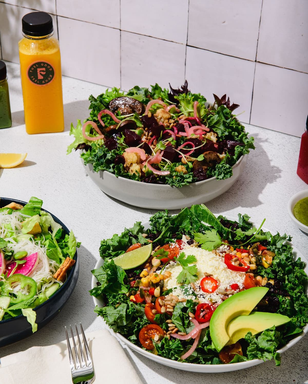 Two bowls of fresh mixed green salads with various toppings including avocado slices, cherry tomatoes, pickled onions, and lime wedges on a white table. Nearby are three bottles of colorful juice or smoothies (yellow, green, and red), a small dish of green dressing, a lemon wedge, and a fork placed on a folded napkin. The background has a tiled wall.