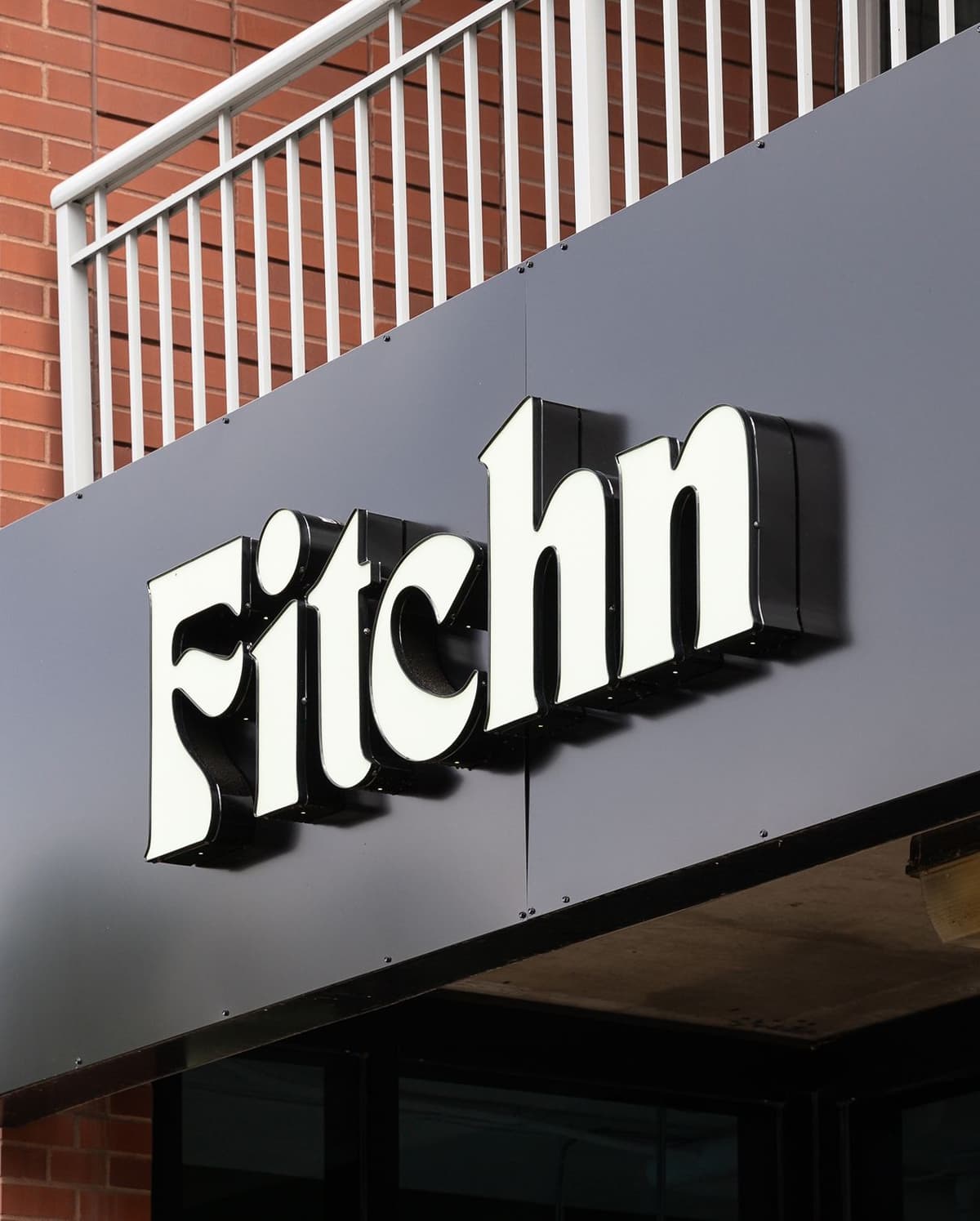 Signage on a black storefront displaying the word "Fitch" in large, white, 3D letters with a brick wall and white railing in the background.