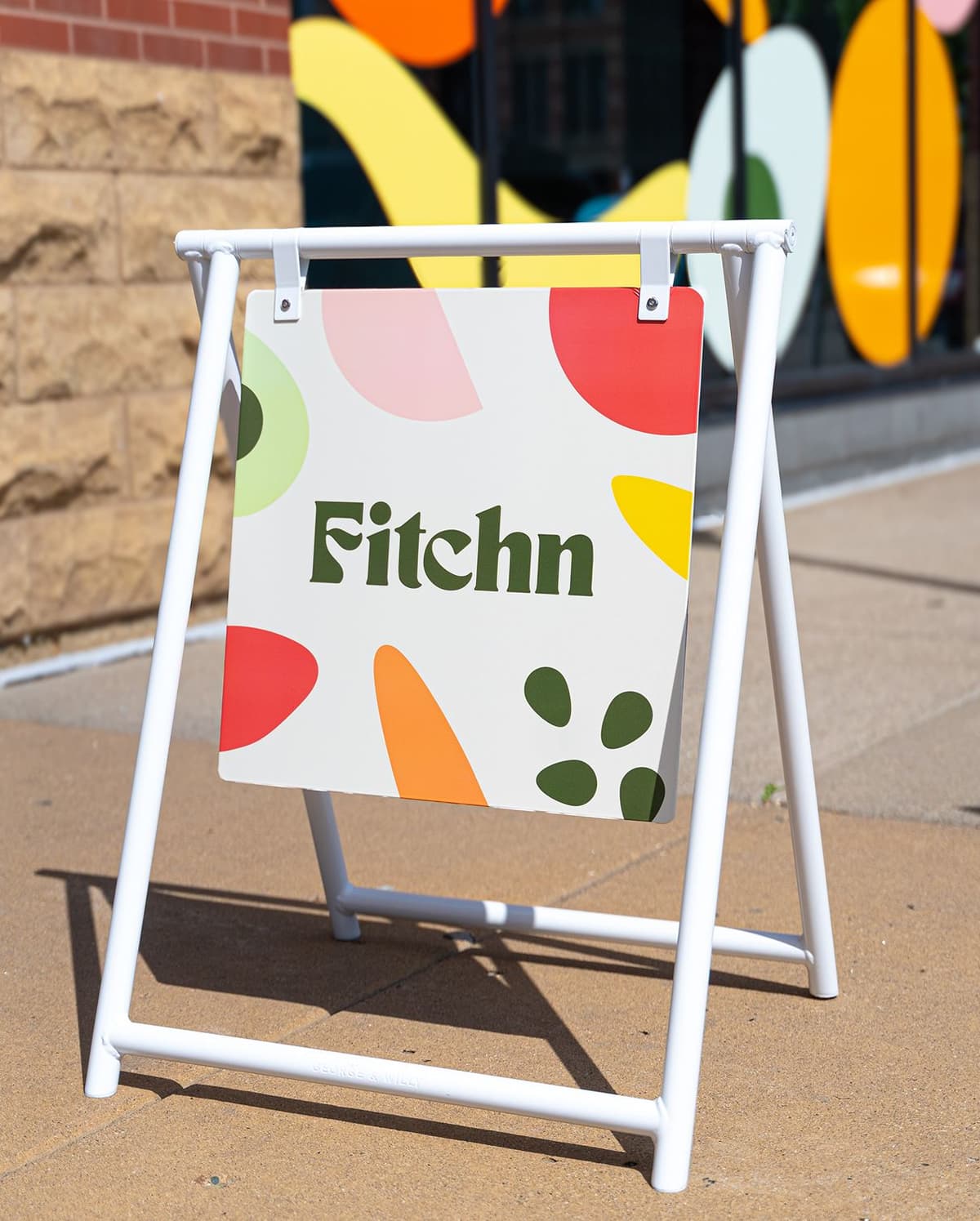 White metal sandwich board sign on a sidewalk with the word "Fitchn" in green text, surrounded by colorful abstract shapes in red, orange, yellow, pink, green, and olive.