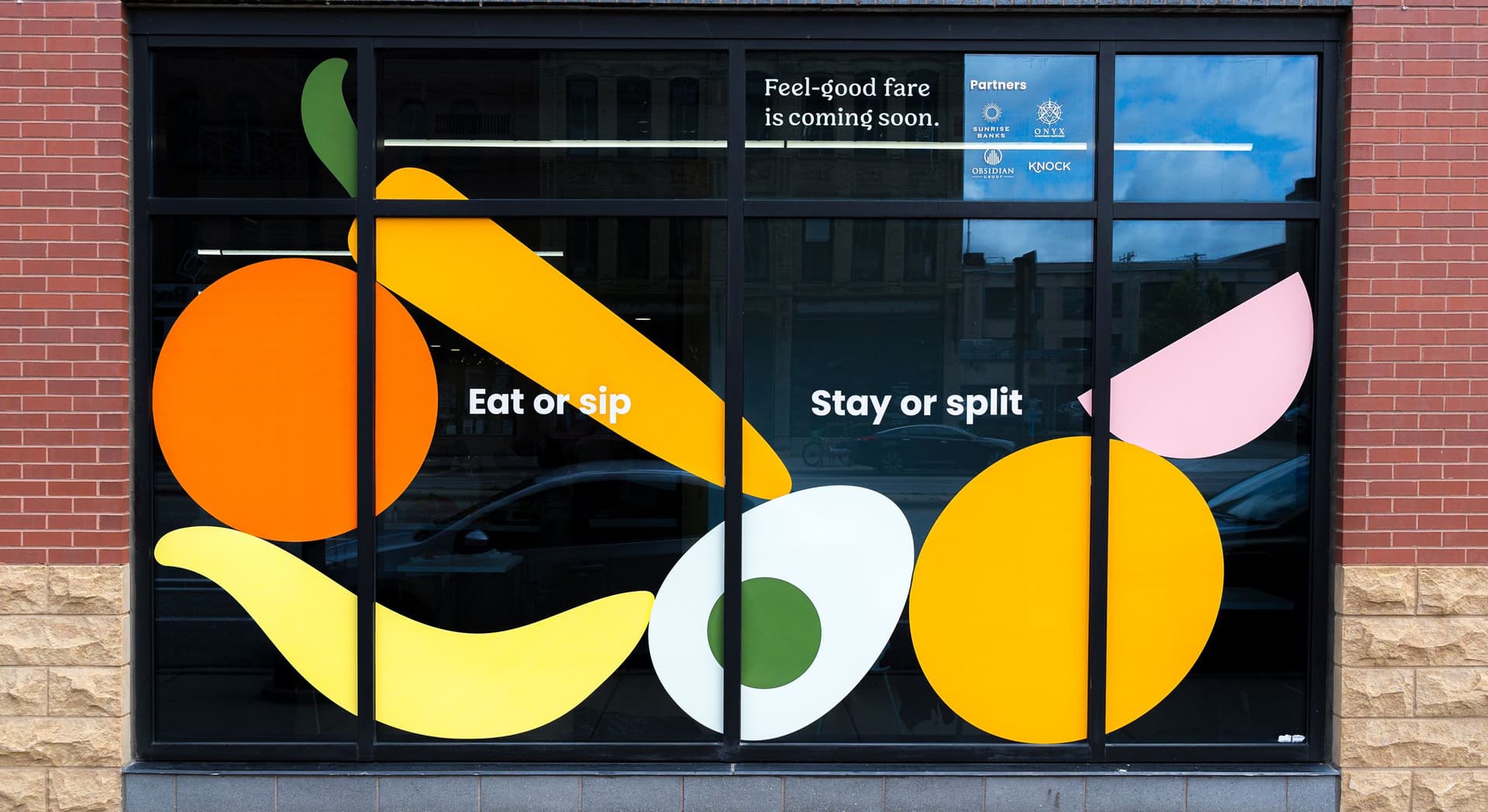 Window display with colorful abstract fruit and vegetable shapes including an orange, banana, avocado, and another circular fruit with text reading "Eat or sip" and "Stay or split." Above, the text reads "Feel-good fare is coming soon" alongside logos of partners.